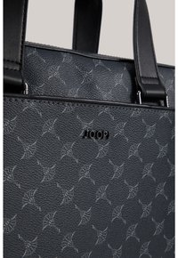 Czarna torba tote o fakturze z wyspecjalizowanego materiału z metalowymi elementami w kolorze srebrnym, z wytłaczanym wzorem i centralnym logo. Podwójne mocne uchwyty do noszenia.