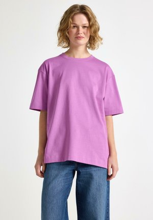 Giovane con capelli biondi corti e mossi, che indossa una maglietta viola oversize e jeans blu, in piedi davanti a uno sfondo bianco uniforme.