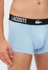 Svetlo modri bombažni bokserci z črnim pasom, na katerem je v belem napisu "LACOSTE" in krokodilovim logotipom. Prikazana oznaka "ULTRA DRY".