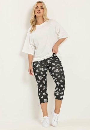 Kvinde iført hvid oversized T-shirt, sorte korte leggings med hvidt margueritprint og hvide sneakers, stående mod en ensfarvet baggrund.