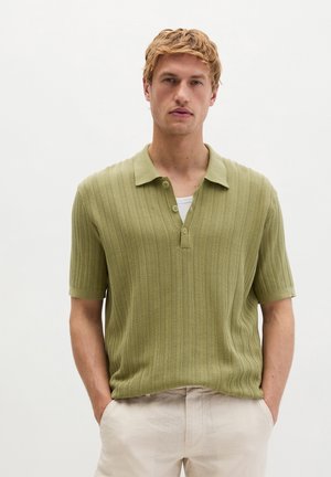 Młody mężczyzna z krótkimi blond włosami, ubrany w jasnozieloną prążkowaną koszulkę polo z krótkim rękawem i beżowe spodnie, ręce w kieszeniach, neutralne tło.