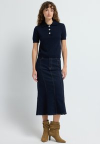 En kvinde med krøllet hår iført en navy kortærmet sweater med perleknapper, en mørk denim midi-nederdel og tan farvede slouchy hæle støvler på hvid baggrund.