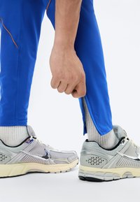 Χέρι που προσαρμόζει το φερμουάρ στο μανσέτι μπλε αθλητικών παντελονιών πάνω από λευκές κάλτσες και γκρι παπούτσια τρεξίματος Nike σε λευκό φόντο.