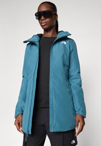Teal vanntett jakke med hette, frontglidelås, elastiske mansjetter, og North Face-logo på venstre bryst. Svart topp under.