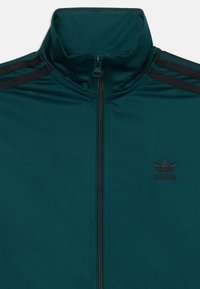 Casaco azul-teal com fecho éclair e gola alta, riscas pretas em destaque e um discreto logo da Adidas preto no peito. Textura de tecido suave.