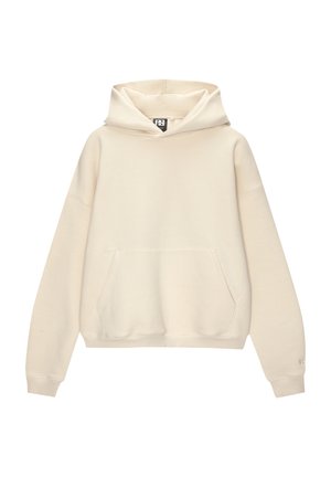 Persoon die een crèmekleurige oversized hoodie draagt met een voorkantzak en losse grijze spijkerbroek, handen in de broekzakken gestopt.