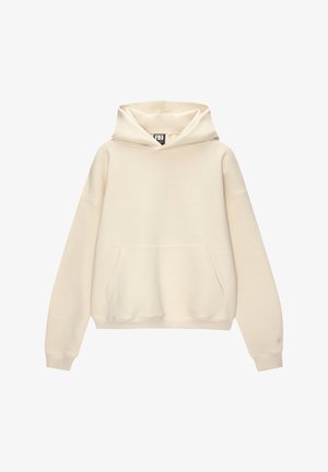 Person trägt einen cremefarbenen oversize Hoodie mit Fronttasche und lockere graue Jeans aus Denim, die Hände in den Hosentaschen.