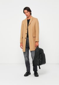 Palton din lână în nuanță bej, cu un design ajustat, tricou negru, blugi gri, rucsac din piele neagră și loafers negri. Stil structurat și modern.
