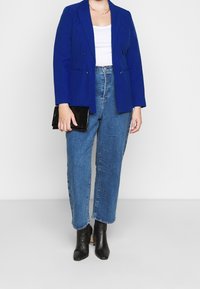 Blazer azul brillante de doble botonadura con solapas muesca, combinado con jeans de cintura alta de color azul claro y botines negros de tacón. Sostiene un clutch negro.