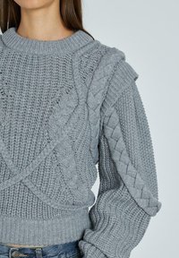 Grauer gestrickter Pullover mit strukturierten Mustern, Kabeldetails und einem breiten Rippensaum. Lange Ärmel mit weiter Passform und überlappendem Design.