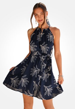 Kobieta ubrana w granatową, bezrękawnikową sukienkę na szyję z białymi wzorami liści, zawiązanym paskiem i wiszącymi kolczykami, pozująca z delikatnym uśmiechem.