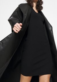 Robe noire à côtes avec décolleté en V, associée à un long manteau en cuir noir. Le manteau présente une texture brillante et des boutons-pression pour la fermeture.