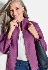 Lila Wildlederjacke mit einem Reißverschluss vorne, getragen über einem weißen Oberteil. Blaue Jeans mit einem Gürtel. Schwarze Handtasche mit einer glatten Textur.