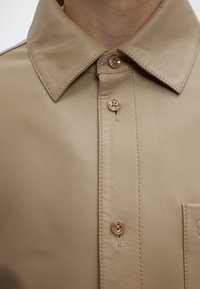 Gros plan d’un col de chemise en cuir beige avec trois boutons fermés et des détails de couture visibles.