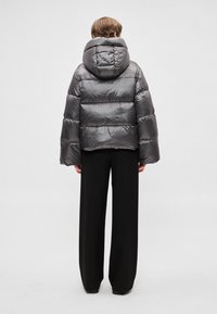 Puffasztott szürke kabát kapucnival, vízszintes varrással és széles ujjal, fekete, laza nadrággal viselve, hátulnézetből.