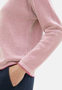 Kötött, hosszú ujjú pulóver, texturált világoskék rózsaszín mintázattal. Bordás mandzsettákkal és szegéllyel rendelkezik, sötétkék nadrág fölött viselik.