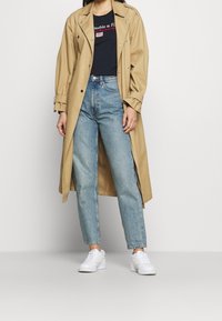 Trench beige sobre una camiseta gráfica azul oscura, combinado con jeans azul claro y zapatillas blancas. Presenta botones y un frente abierto.