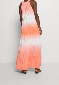 Mujer vistiendo un vestido maxi sin mangas, de volantes y con un patrón ombre en tonos de rosa, blanco y coral, complementado con sandalias color crema.