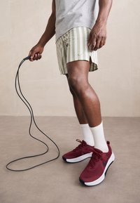 Red athletic shoes with black accents, gray t-shirt, and green-striped shorts. Holding a jump rope, wearing white socks. Smooth background.