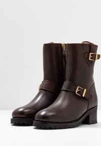 Hnědé kožené kotníkové boty s kulatou špičkou, zlatými sponami a akcenty, postranním zipem a strukturovanou gumovou podrážkou. Design s blokovým podpatkem.