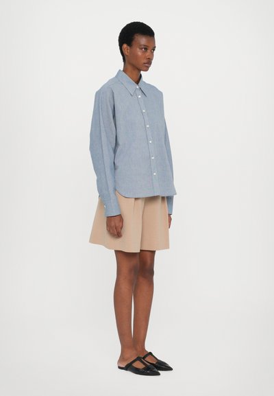 Blaues Chambray-Hemd mit spitzem Kragen, durchgehender Knopfleiste und langen Ärmeln, kombiniert mit einem beigen A-Linien-Rock und schwarzen Ballerinas.