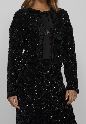 Czarny, długi rękaw top i spódnica z cekinami, z dwoma czarnymi kokardami na piersi, noszone przez kobietę na jednolitym tle.