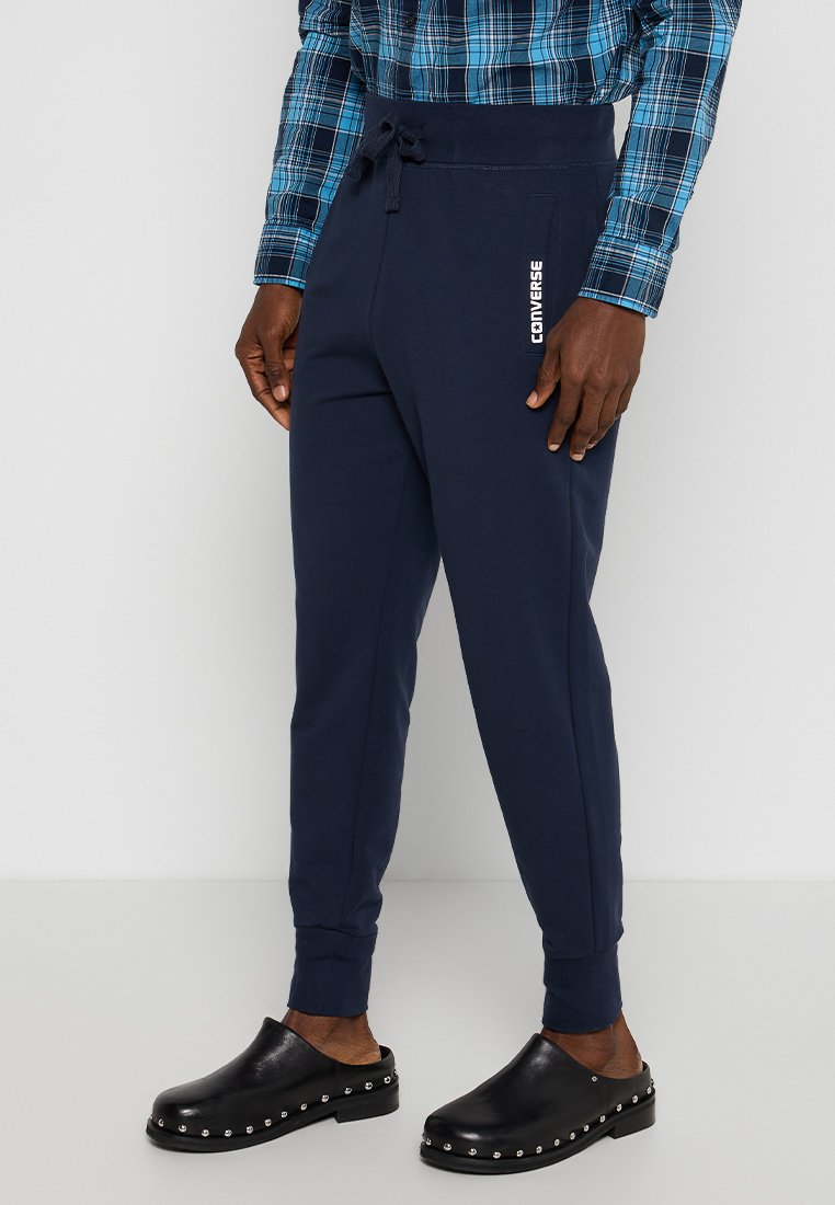 Pantaloni jogger blu navy con vita elastica e laccetto, dotati di tasca laterale e logo 'Converse' stampato, abbinati a scarpe slip-on nere.
