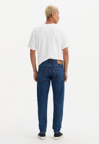 Moški s kratkimi blond lasmi, oblečen v belo majico, modre kavbojke in črne teniske, stoji obrnjen stran od bele ozadja.