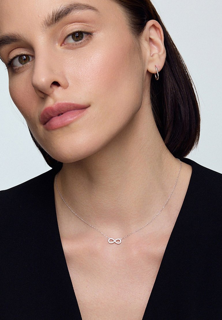 Srebrny naszyjnik w kształcie nieskończoności na delikatnym łańcuszku, z gładkim, wypolerowanym wykończeniem. Modelka w czarnej bluzce z dekoltem w serek.