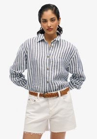 Camisa de lino a rayas azules y blancas con diseño de botones; combinada con pantalones cortos blancos deshilachados y un cinturón trenzado marrón.