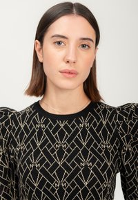 Giovane donna con capelli castani lisci, che indossa un maglione nero con un motivo geometrico beige e spalle a sbuffo, su uno sfondo uniforme.