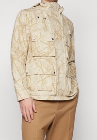 Beige jacka med ett kartliknande mönster, flera framlommor, hög krage och tryckknappar. Tillverkad av lättviktsmaterial.