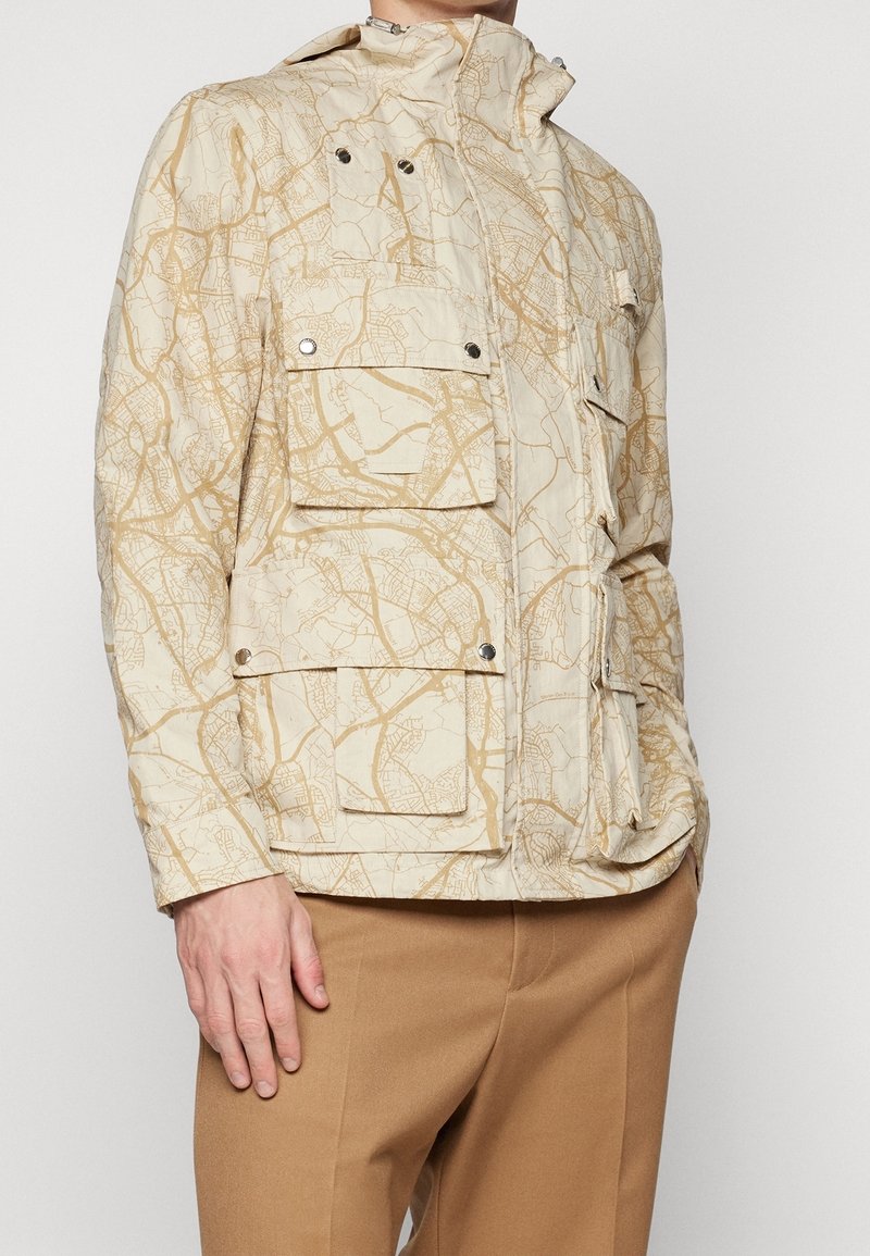 Beige jacka med ett kartliknande mönster, flera framlommor, hög krage och tryckknappar. Tillverkad av lättviktsmaterial.