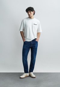 White short-sleeve t-shirt with black text. Slim-fit dark blue jeans and light-colored sneakers with brown accents. Minimalist design.