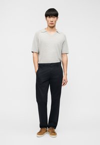 Homme portant une chemise à manches courtes en maille gris clair, un pantalon noir et des baskets marron, debout avec une main dans la poche devant un fond uni.