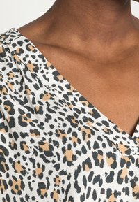 Blusa con stampa leopardata, caratterizzata da uno sfondo bianco con macchie nere e beige, tessuto morbido e scollatura a V. Dettaglio di drappeggio sobrio sulla spalla.