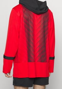 Roter langärmeliger Hoodie mit einer schwarzen Kapuze, der ein strukturiertes Zickzackmuster und schwarze Streifen an den Ärmeln aufweist. Das Material erscheint leicht.