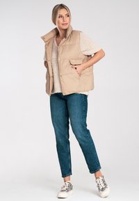 Beżowa kamizelka puchowa z wysokim kołnierzem, zapięciem na napy i dwoma przednimi kieszeniami, noszona na kremowym swetrze robionym na drutach i niebieskich dżinsach. Dodatkowo casualowe obuwie.