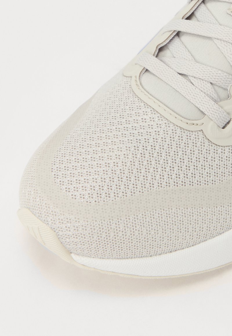 Zapatilla deportiva de color gris claro con parte superior de malla transpirable, puntera redondeada y entresuela suave. Presenta cordones planos y patrones de textura sutiles.