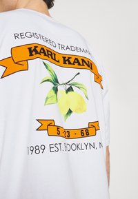 Bílý tričko s potiskem žlutého citronu, oranžovou stuhou s textem "KARL KANI" a detaily o značce a datu založení.