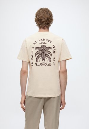 Personne aux cheveux bouclés portant un t-shirt beige affichant un graphique de palmier et le texte français « La famille et l'amour avant tout » au dos.