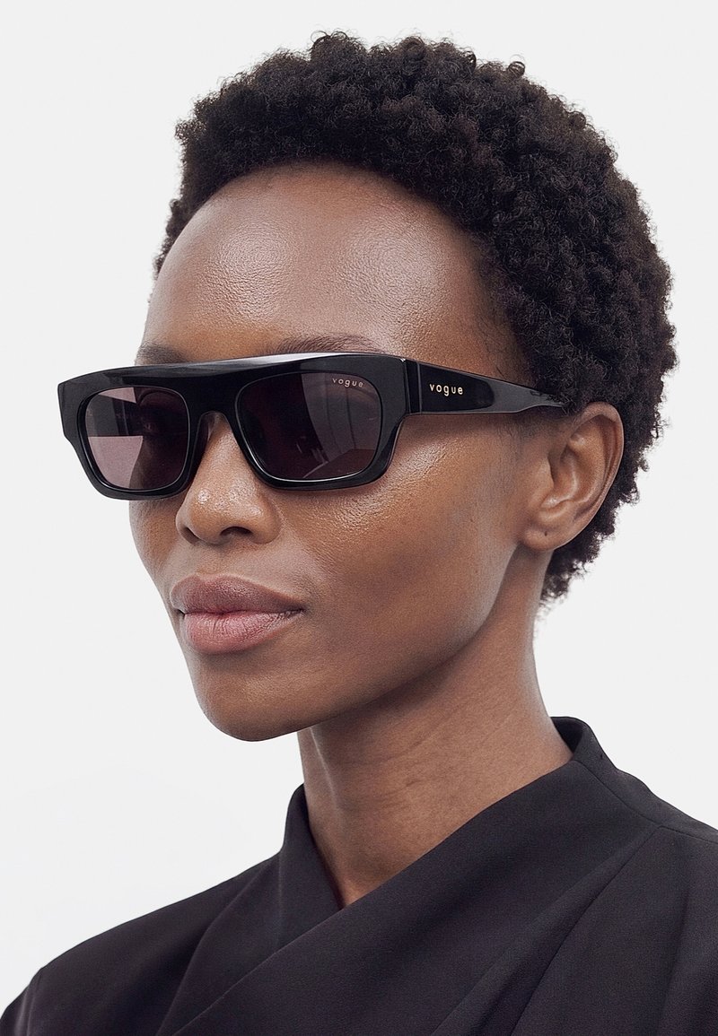 Personne avec des cheveux courts et bouclés portant des lunettes de soleil rectangulaires noires Vogue et un haut noir à col montant, légèrement tournée vers la droite.