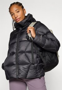 Sieviete ar pītām matu šķipsnām smaidot valkā melnu pūkainu jaku un tur rokās mugursomu, fona attēls ir neitrāls.