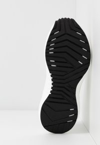 Idrottssko med en svart gummisula med struktur, som har zigzagmönster och ribbningar, samt en vit överkant för stabilitet.