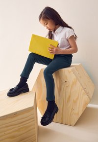 Egy gyermek fehér rövid ujjú inget, zöld nadrágot és fekete mokaszint visel, miközben sárga tabletet tart a kezében, és fából készült geometriai formákon ül.