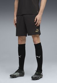 Sorte fodboldshorts med gyldent "KING" tekst; sorte strømper med hvid Puma-logo; sorte og grønne fodboldstøvler med tekstureret design.