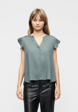 Młoda kobieta ubrana w szarą, bezrękawnikową bluzkę z guzikami i marszczonymi ramionami oraz czarne skórzane spodnie, stojąca na jednolitym tle.