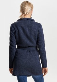Cappotto avvolgente in misto lana blu navy con collo, maniche lunghe, vita con cintura e finitura testurizzata. Presenta una lunghezza fino a metà coscia e una silhouette elegante.