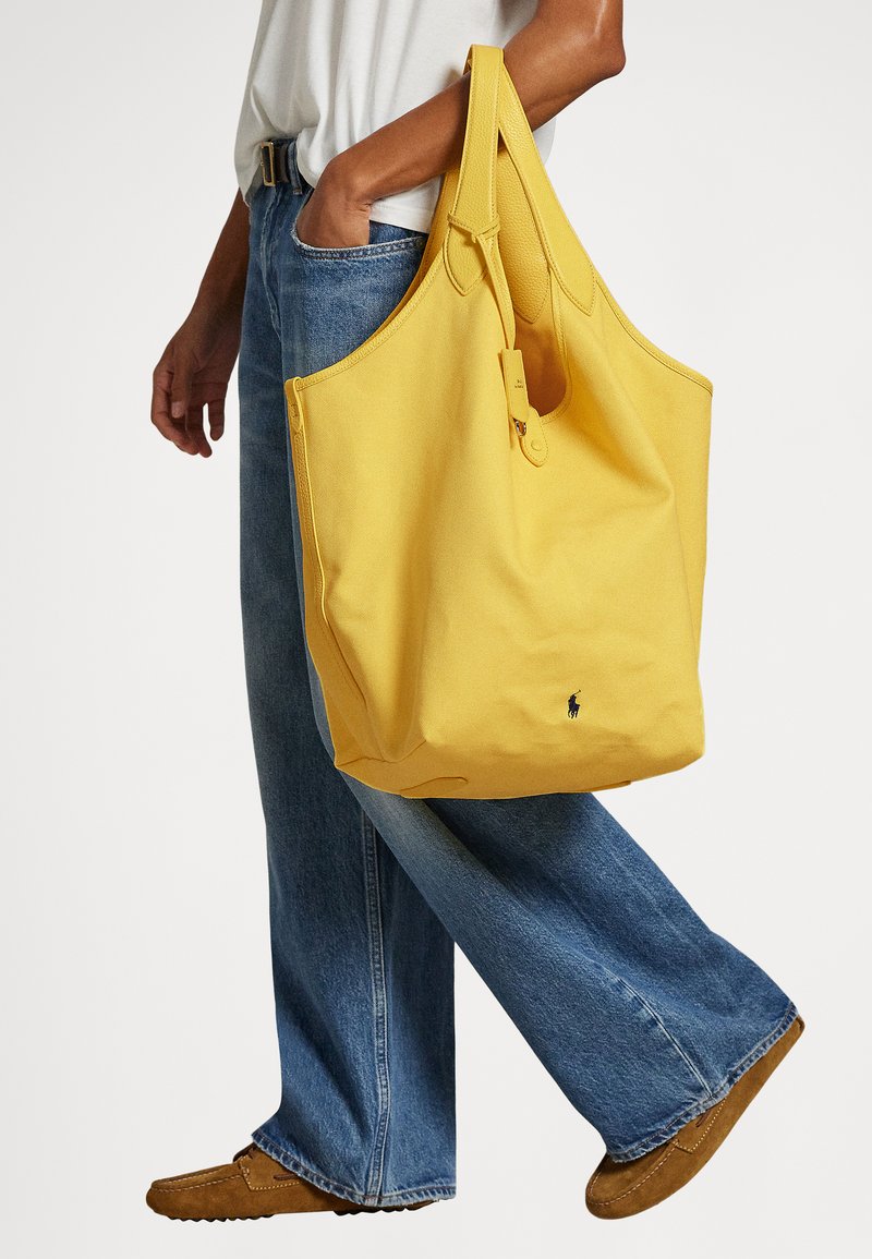 Yellow fabric tote bag with a smooth texture, wide opening, and a small logo. Worn on shoulder, accompanied by loose blue jeans and brown shoes.