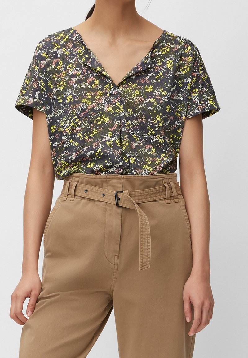 Dunkelbluse mit kurzen Ärmeln im floralen Design, verziert mit leuchtend gelben, pinken und weißen Blumen auf einem dunklen Hintergrund, kombiniert mit beigen, taillierten Hosen.